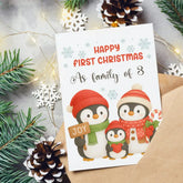Christmas card with penguins and text 'Happy First Christmas as family of 3' surrounded by festive decorations.