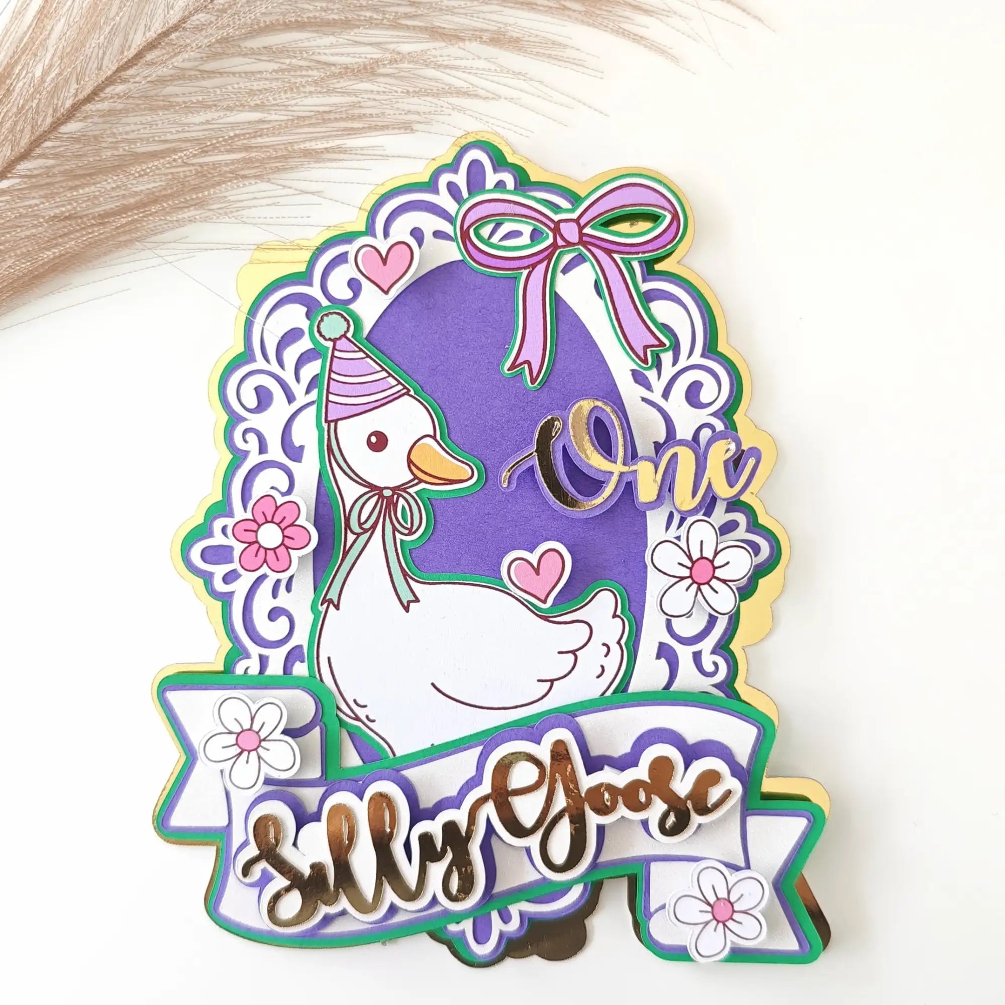 A purple and white layered paper cutout featuring a cartoon goose wearing a party hat, adorned with floral details and the words ’one’ and ’silly goose.’