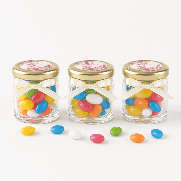 Three clear glass jars filled with colorful jelly beans, each tied with a white ribbon and topped with a gold lid, arranged on a white background with a few loose jelly beans in front.