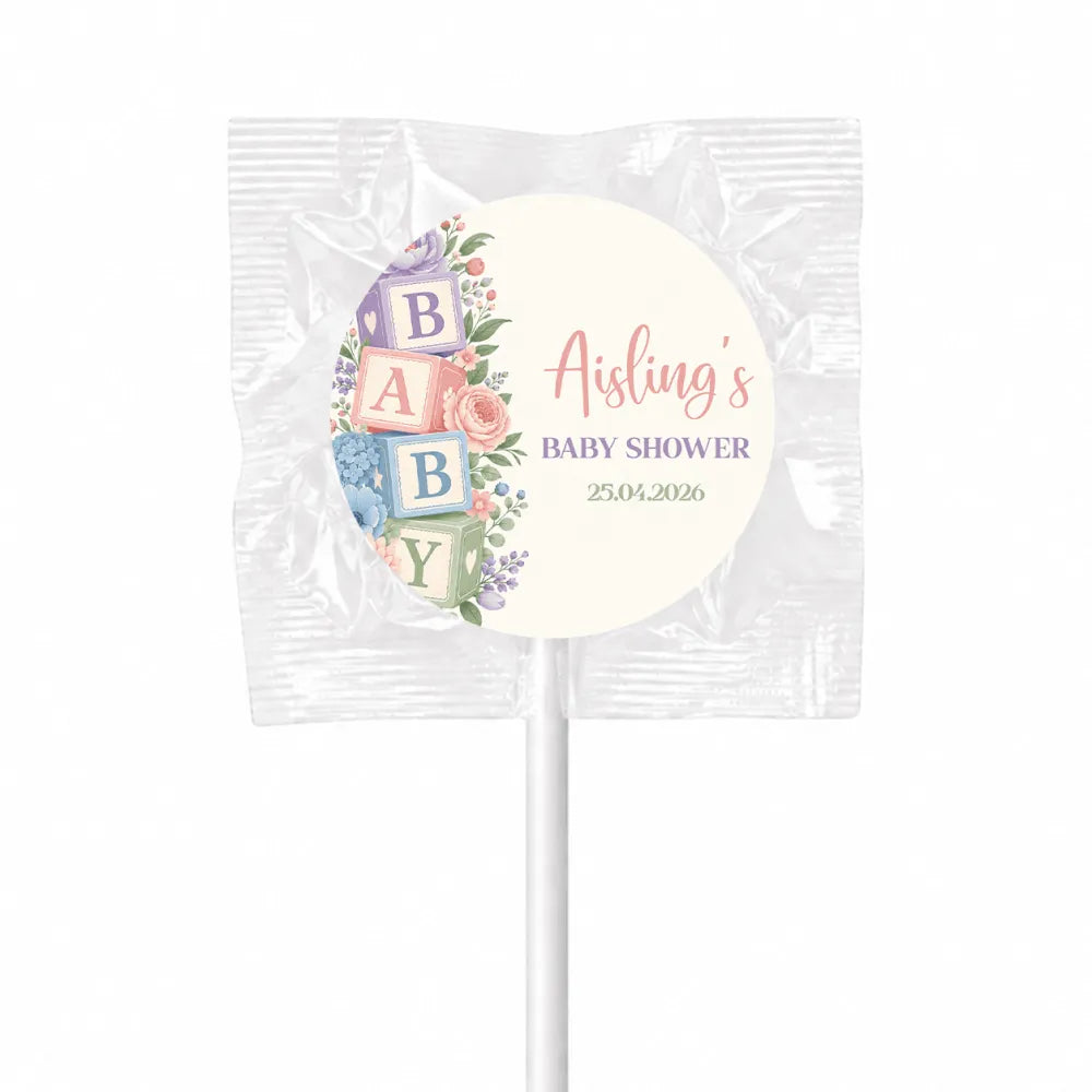 A pastel baby shower lollipop favour wrapped in clear foil with a circular label featuring alphabet blocks shower design