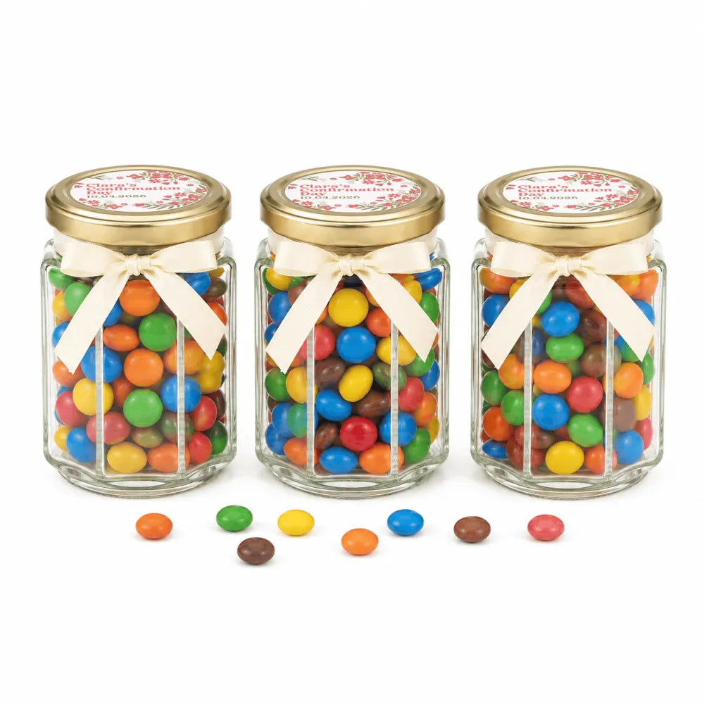 Three clear glass hex jars with 48mm pale gold lids and personalised sticker printed ribbons filled with colorful m&m’s for Blossom Grace Confirmation favors