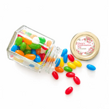 A clear glass hex jar tipped over with colorful jelly beans and a 48mm pale gold twist lid with a personalised sticker printed
