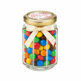 A clear hexagonal glass jar with a pale gold twist lid and cream ribbon bow filled with colorful candies and a personalised sticker printed blossom grace confirmation favour