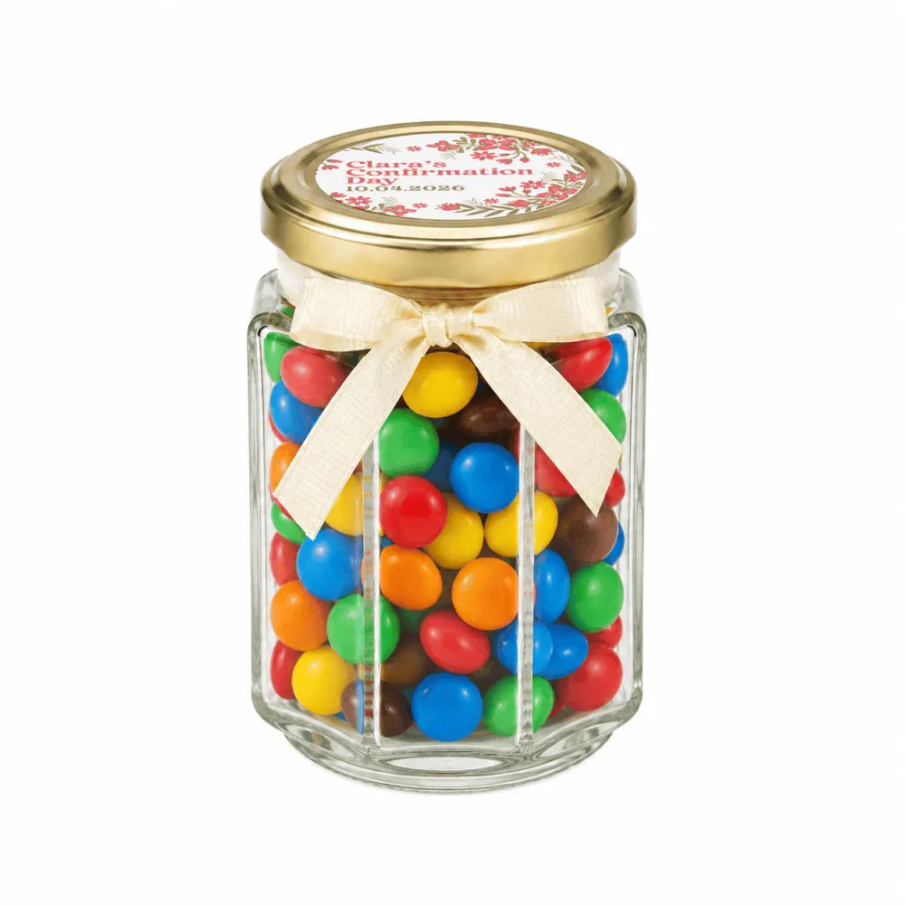 A clear hexagonal glass jar with a pale gold twist lid and cream ribbon bow filled with colorful candies and a personalised sticker printed blossom grace confirmation favour
