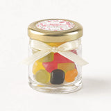 A clear glass mini jar favour with 43mm pale gold twist lid cream ribbon bow and personalised sticker printed label filled with gummies
