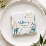 A white chocolate bar loose wrapped in silver foil with blue gold floral patterns and christening details