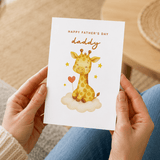 Person holding a Father's Day card with a giraffe illustration on a wooden floor.