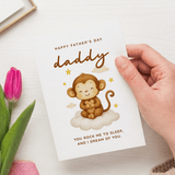 Hand holding a Father's Day card with a monkey illustration and text, next to a pink tulip.