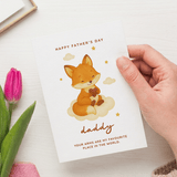 Hand holding a Father's Day card with a fox illustration and pink tulip on a white surface.