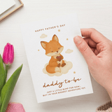 Hand holding a Father's Day card with a fox illustration and pink tulip on a white surface.