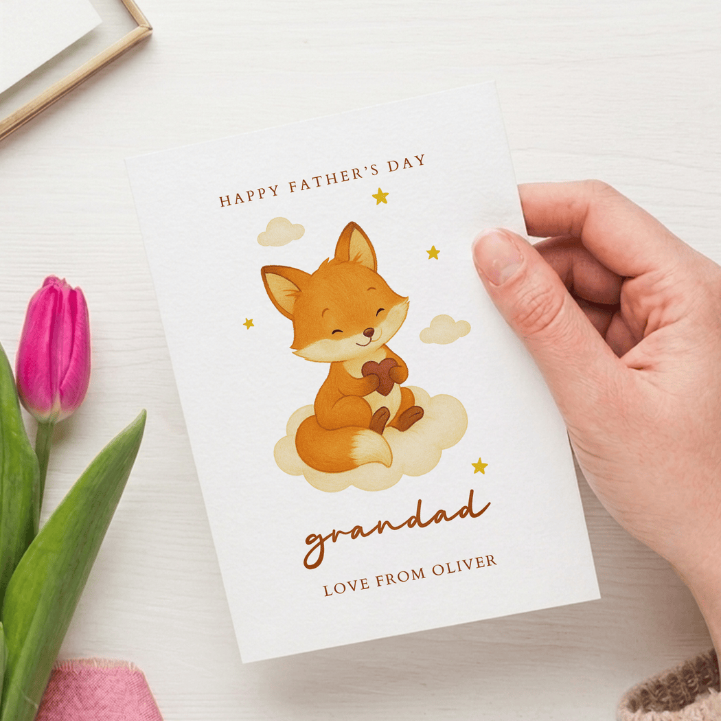 Hand holding a Father's Day card with a fox illustration and text, next to pink tulips on a light surface.