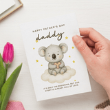 Hand holding a Father's Day card with a koala illustration and pink tulip on a white surface.