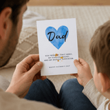 Child holding a Father's Day card with a blue heart and text, sitting on a couch with an adult.