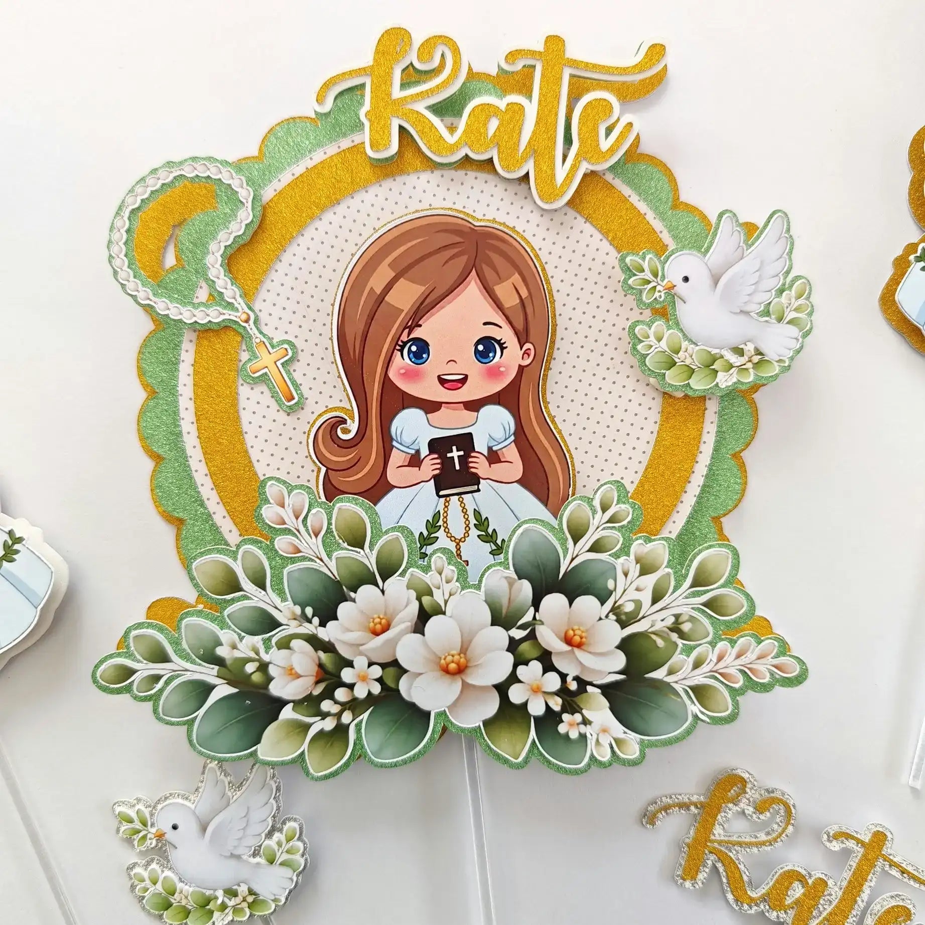 A decorative cake topper featuring a cartoon girl with brown hair and blue eyes, holding a black book, surrounded by white flowers, green leaves, two doves, and the name ’kate’ in gold script.