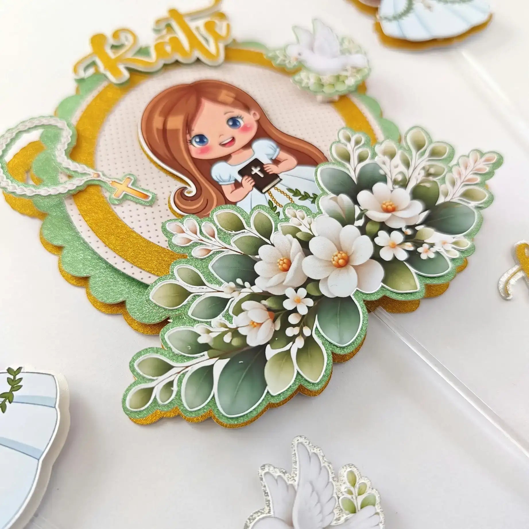 A decorative, layered cake topper featuring a cartoon girl with long brown hair and blue eyes holding a small black book with a cross emblem, surrounded by white flowers and green foliage on a glittery gold and light green base.