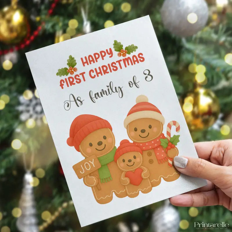 Greeting card with gingerbread family illustration and text 'Happy First Christmas as family of 3' held in front of a decorated Christmas tree.