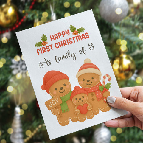 Greeting card with gingerbread family illustration and text 'Happy First Christmas as family of 3' held in front of a decorated Christmas tree.