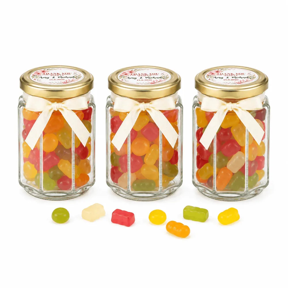 Three hexagonal glass jars with gold lids and cream ribbons filled with gummy candies for floral wreath wedding favours