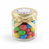 A clear glass mini jar wedding favour filled with colorful candy and topped with a gold lid and ribbon bow floral wreath design