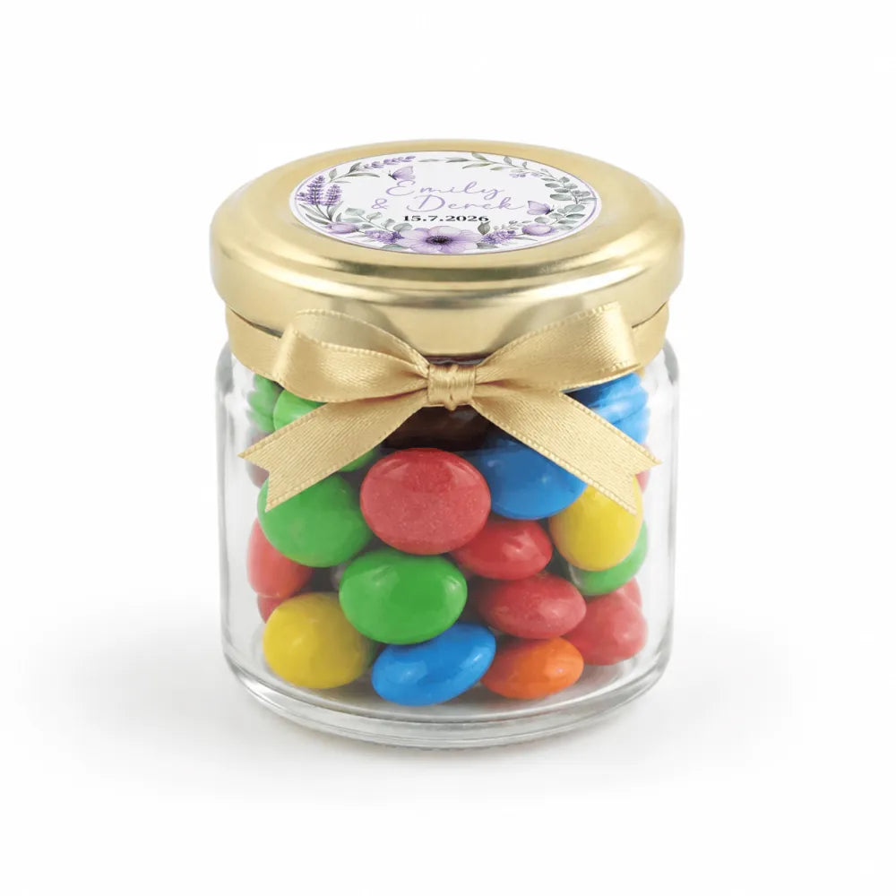 A clear glass mini jar wedding favour filled with colorful candy and topped with a gold lid and ribbon bow floral wreath design