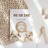 Gender reveal card with two baby bunnies and text 'He or She' on a textured surface.