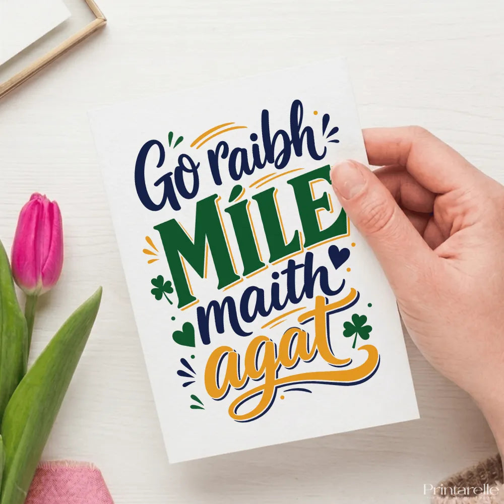 A white greeting card with go raibh míle maith agat calligraphy in navy blue green and gold with shamrocks