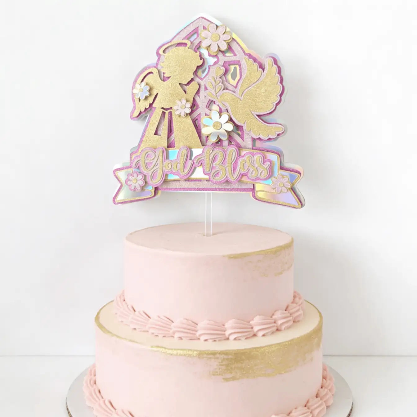 A delicate pink and gold cake topper featuring an angel, a dove, and the phrase ’god bless’ in glittery script.