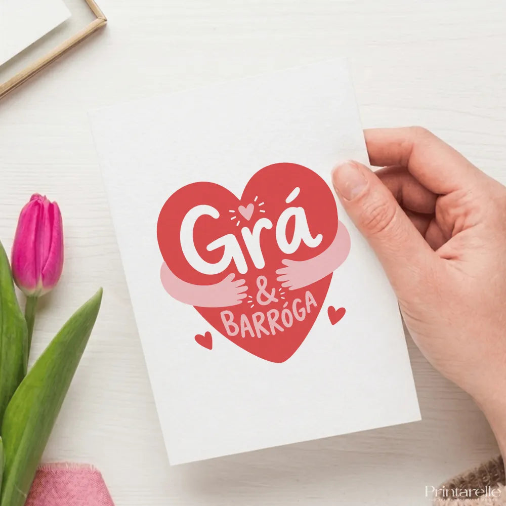 A white greeting card featuring a vibrant red heart with playful white grá agus barróga script and pink hugging arms