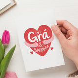 A white greeting card featuring a vibrant red heart with playful white grá agus barróga script and pink hugging arms
