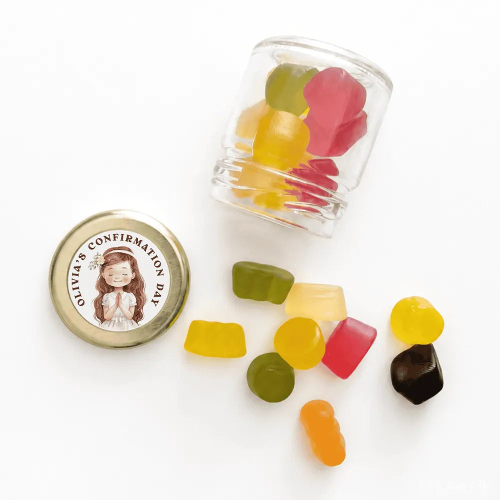 A clear glass mini jar favour with a 43mm pale gold twist lid and personalised sticker printed for confirmation day spilling colorful gummies