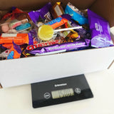 A white cardboard late late toy show sweet box filled with colorful candy wrappers and a yellow lollipop