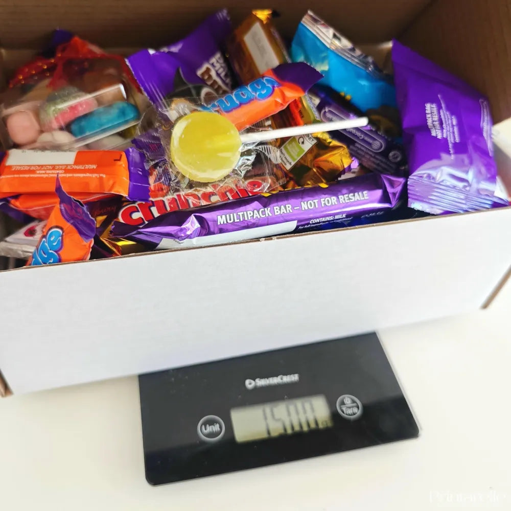 A white cardboard late late toy show sweet box filled with colorful candy wrappers and a yellow lollipop