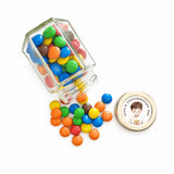 A clear glass hex jar tipped over spilling colorful mms with a 48mm pale gold twist personalised sticker printed label