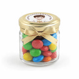 A clear glass mini jar favour filled with colorful candy, featuring a pale gold twist lid and personalised sticker printed label