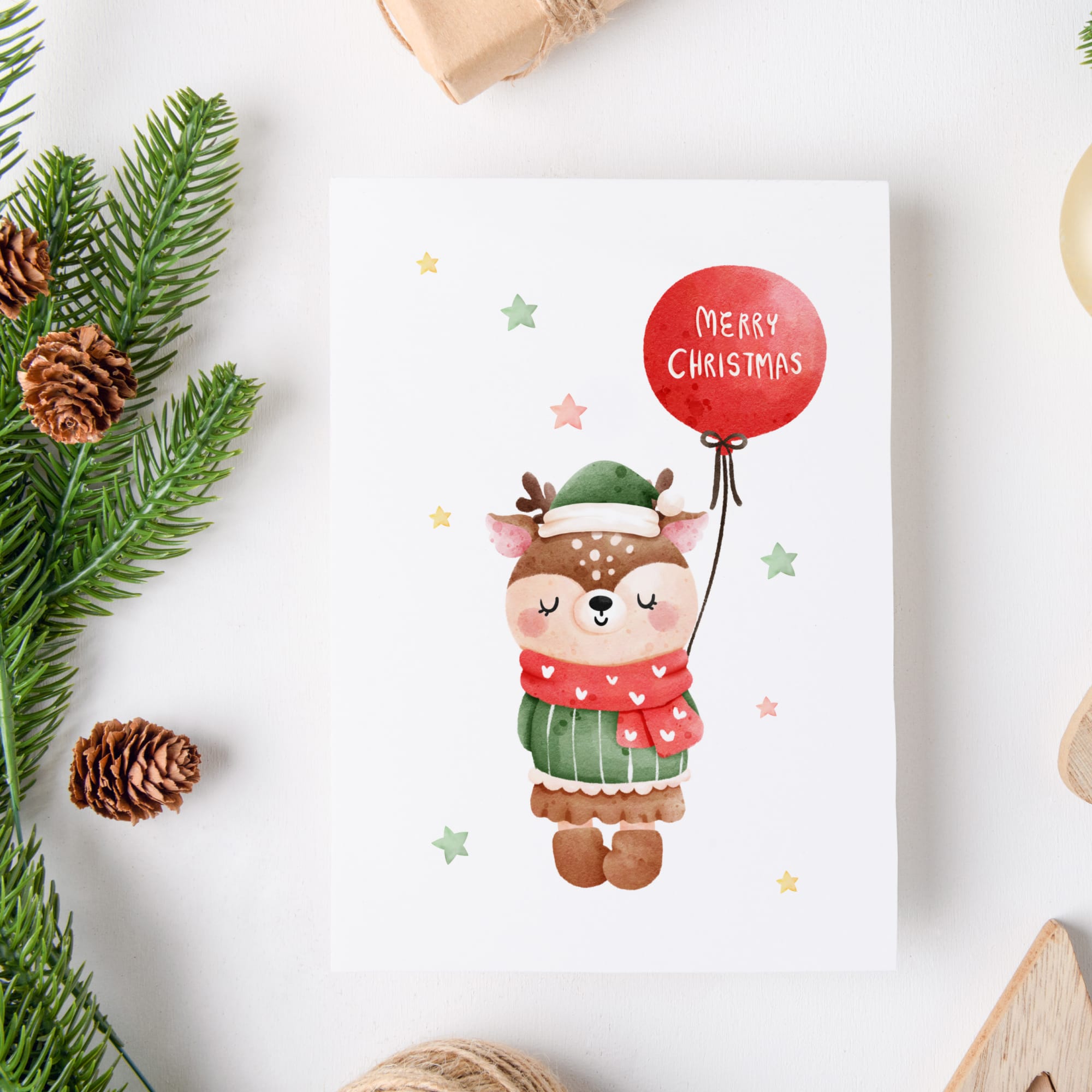 Christmas card with a cartoon deer holding a red balloon with 'Merry Christmas' text, surrounded by pine branches and pinecones on a white background.