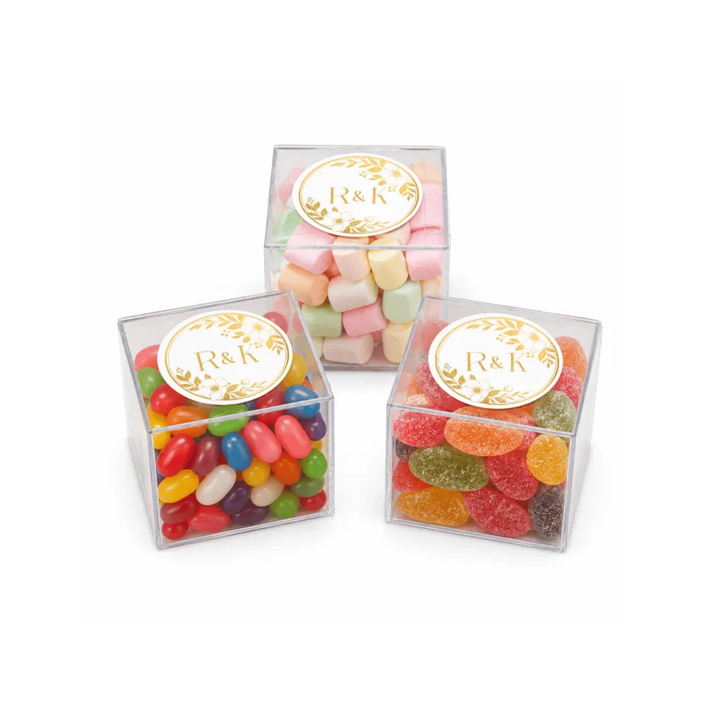Three clear boxes filled with colorful candies sealed with gold embossed r&k label featuring a monogram wreath for wedding date