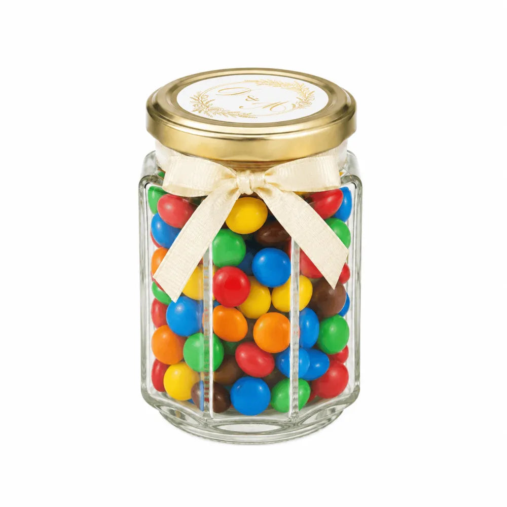 A clear hexagonal glass jar with a gold lid and cream ribbon filled with colorful mms featuring a monogram wreath wedding favour design