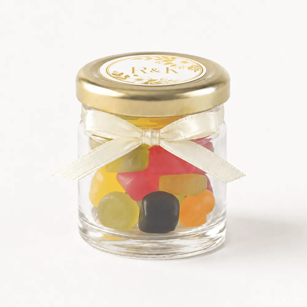 A clear glass jar with gold lid and cream ribbon bow filled with colorful gummies for monogram wreath wedding favours