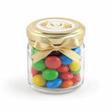 A clear glass jar with a gold lid and ribbon bow filled with colorful m&m’s wedding favours featuring a monogram wreath foil sticker