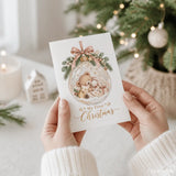A white christmas card featuring a glass bauble with a sleeping teddy and bunny tied with a soft pink ribbon bow