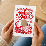 A festive white nollaig shona card with red foil holiday illustrations in irish language and a circular cutout