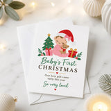A white 300gsm christmas card showing a cartoon baby in pink pajamas with santa hat holding a teddy bear by gifts