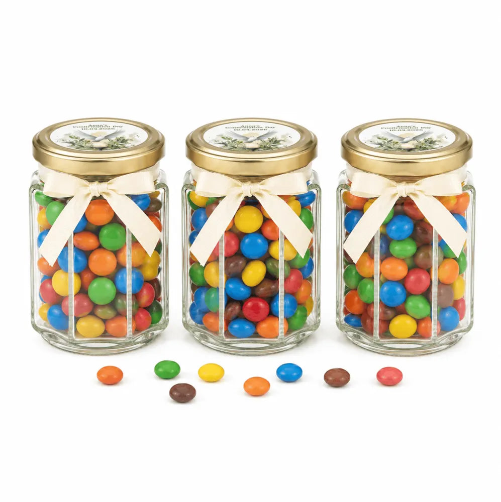 Three clear glass jars with pale gold twist lids and cream ribbons filled with colorful m&ms for peaceful dove confirmation favor
