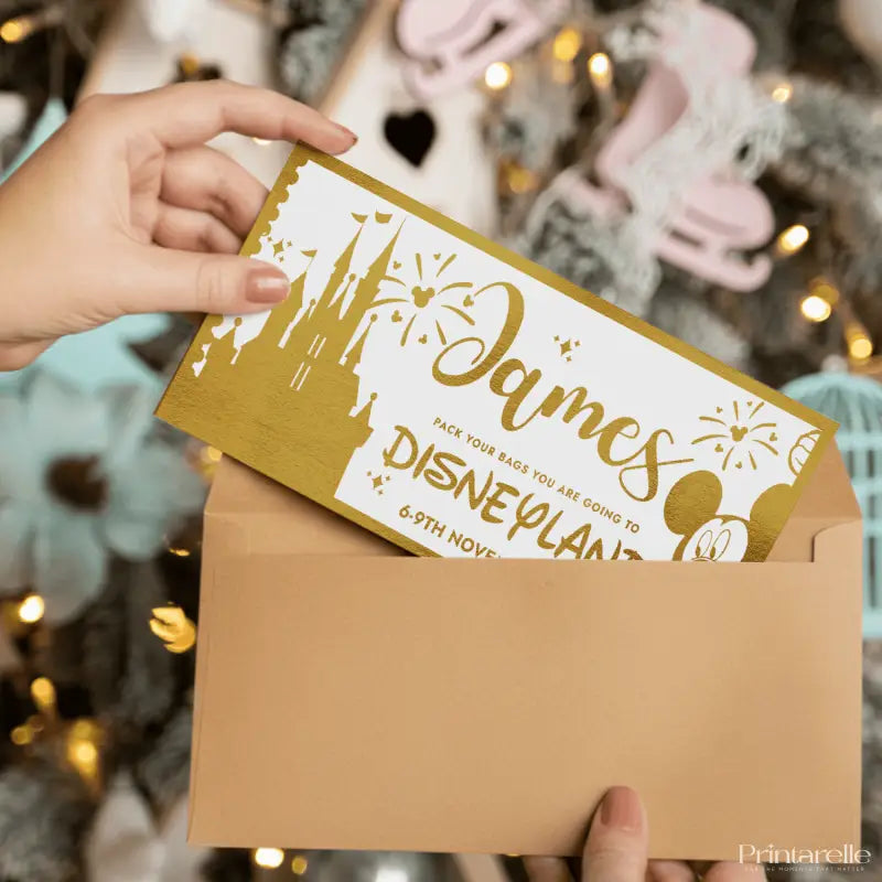 Hand holding a Disney-themed invitation card with 'James' and 'Disneyland' text, against a festive background.