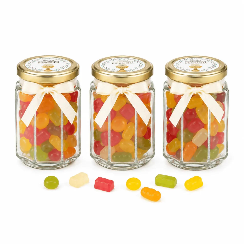 Three hexagonal glass jars with pale gold twist lids and cream bows filled with gummy sweets with personalised sticker