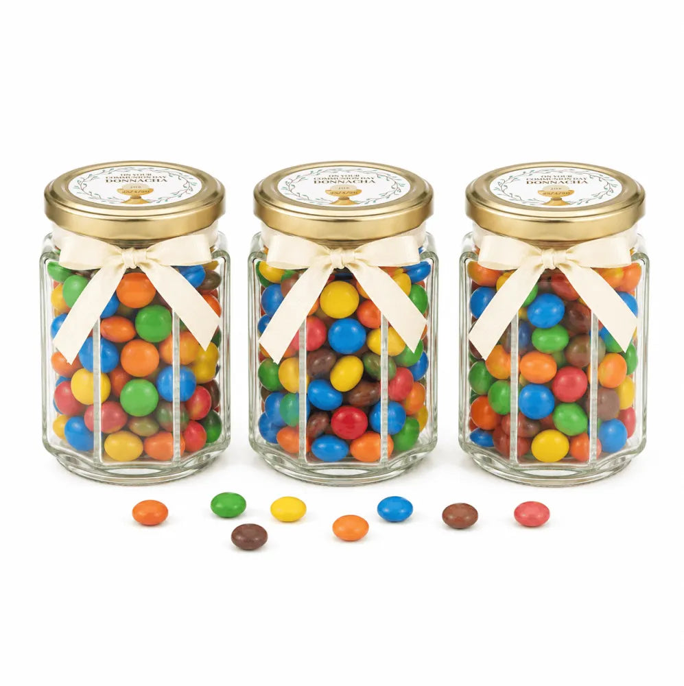 Three clear 106ml glass hex jars with gold lids and cream ribbons filled with colorful m&m’s and personalised sticker