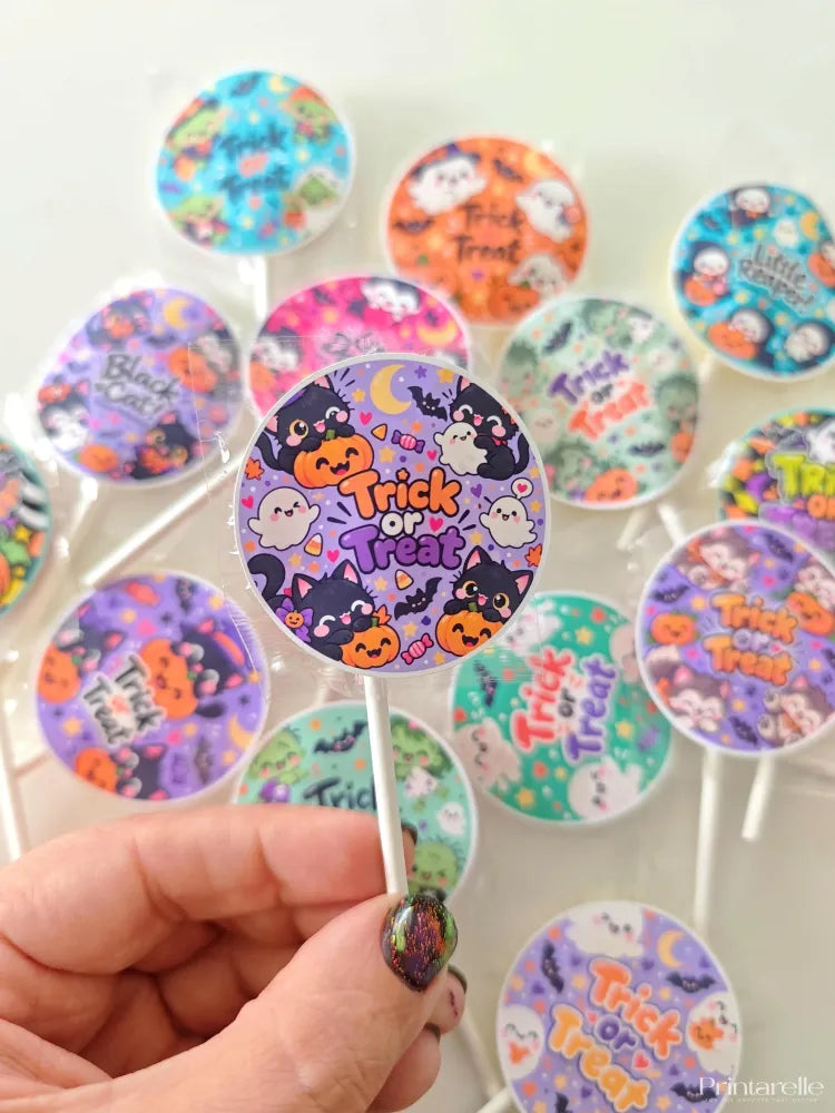 A hand holds a round halloween lollipop with colorful cartoon black cat pumpkins on assorted fruit lollipops candy surface