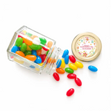 A clear glass hex jar tipped over spilling colorful jelly beans with a 48mm pale gold twist lid and personalised sticker printed