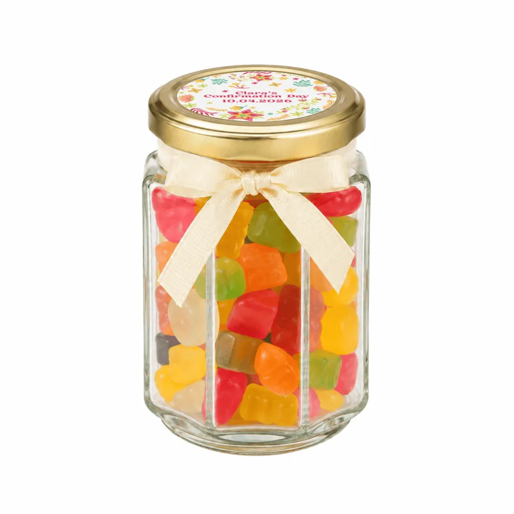 A clear glass personalised hex jar favour with a 48mm pale gold twist lid and cream ribbon bow filled with gummy bears