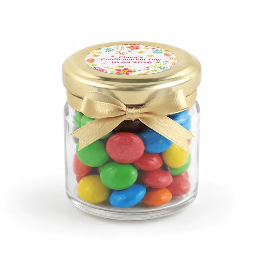 A clear glass jar favour filled with colorful m&m’s in a 43mm pale gold twist lid with a personalised sticker printed